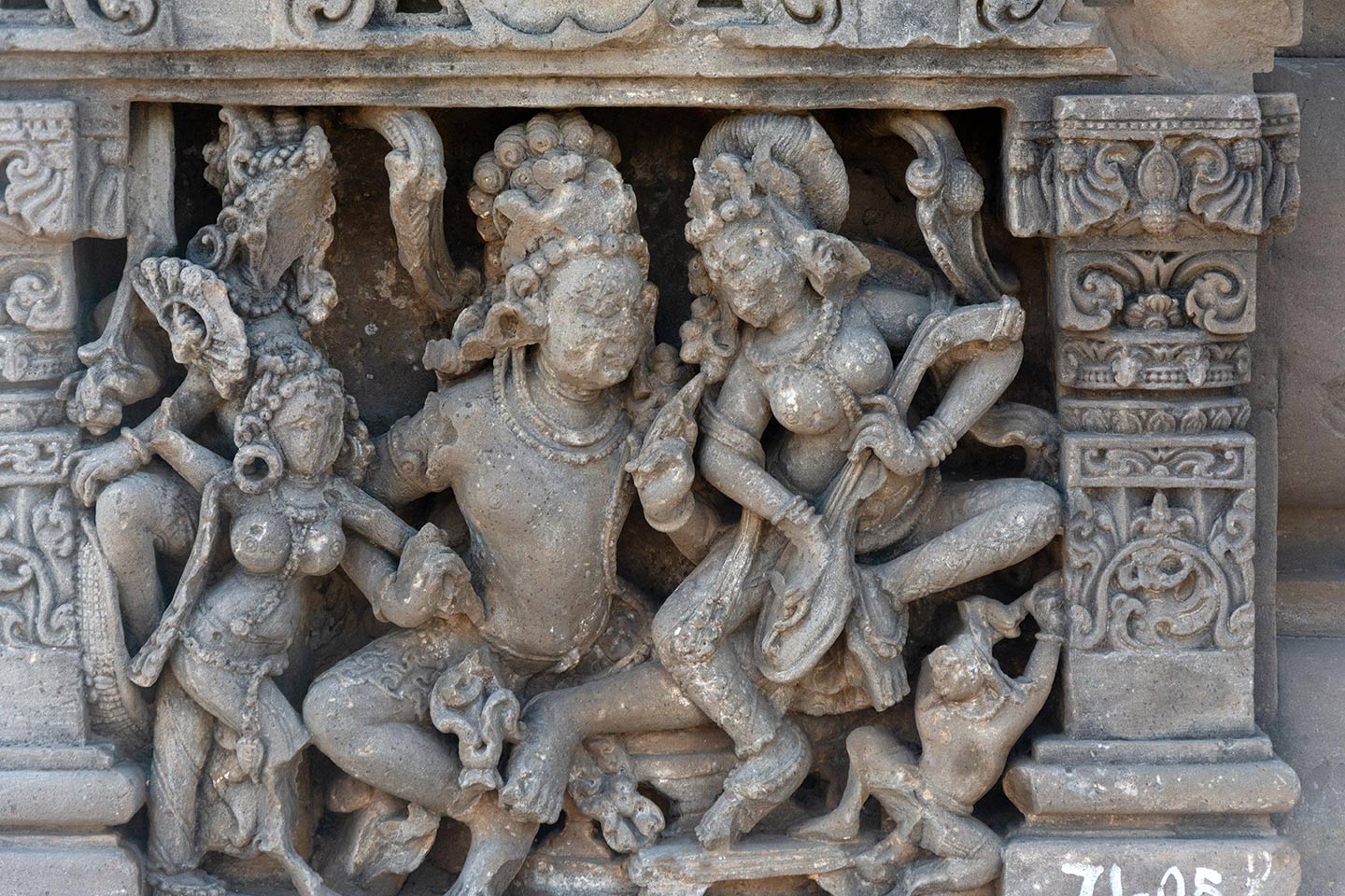 Image 7: A central male figure sits in a lalitasana pose on a raised seat, gazing towards a central female figure who is playing a stringed musical instrument on his left thigh. The female figure is also in a lalitasana pose, and her right foot is resting on a footrest. She has elevated her body slightly to the left of the man. Hence, she is looking down towards the man. Positioned below her left leg is the figure of a male attendant. With his right hand, the central male figure forcefully pulls a female attendant (holding a hand fan) towards him.