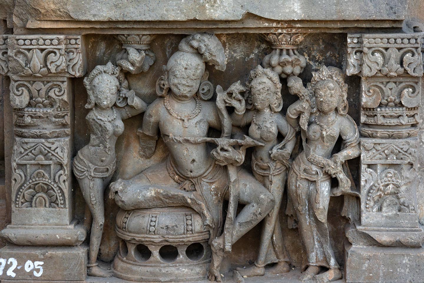 Image 8: The central male figure sits on a circular raised seat in a lalitasana pose, firing an arrow from a bow. Standing on his left are two female figures (faces defaced), looking towards him. The female on his immediate left is holding a flower in her right hand. The female to his far left is holding a floral scroll. The female figure on his right has sustained damage. A bunch of mangoes hang above them. Mangoes are symbols of kama, or sexual desire; this may be a depiction of Kamadeva (the Hindu god of erotic love, pleasure, and desire), and the females may be apsaras (celestial nymphs) or his consort, Goddess Rati.