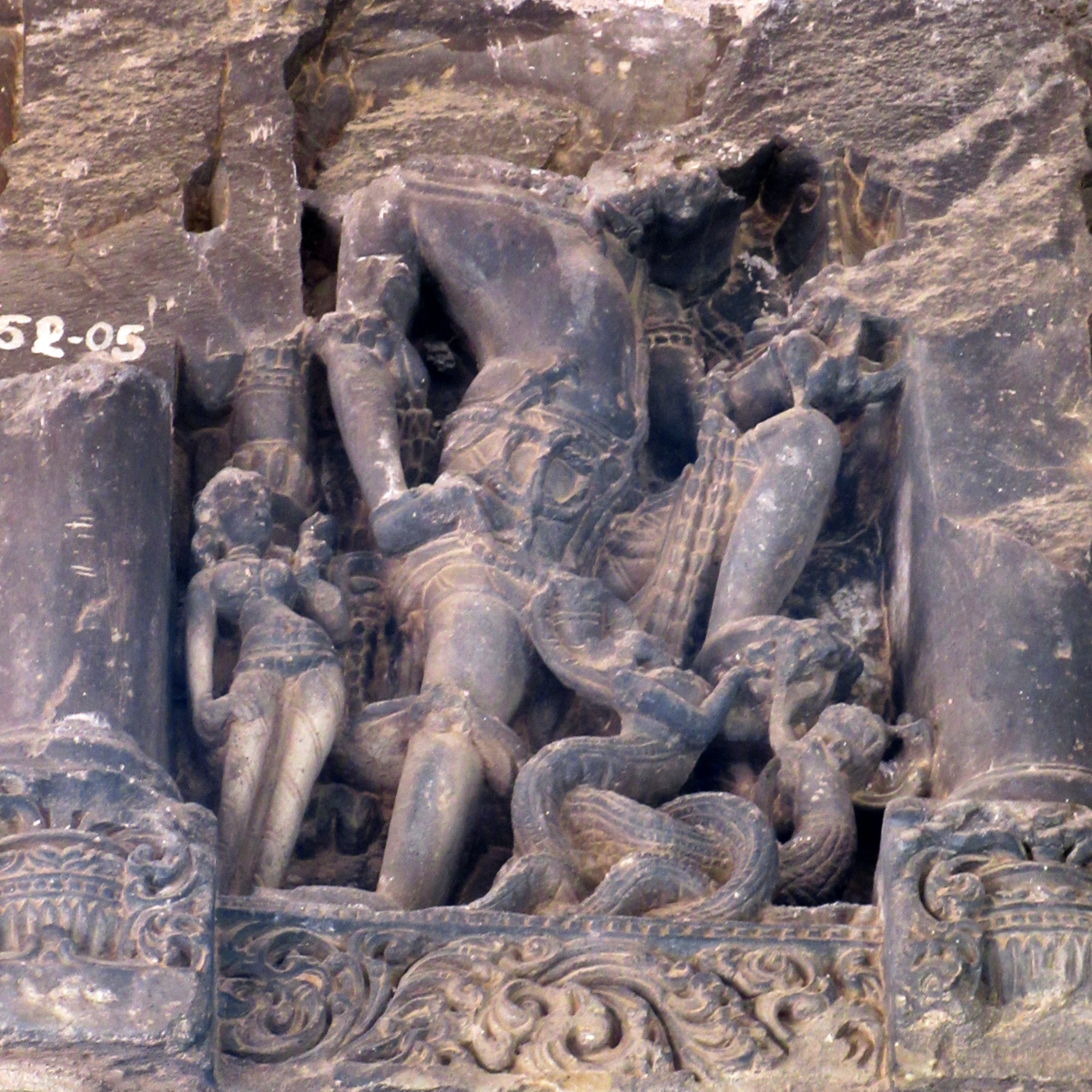 Image 12: A four-armed male deity firmly stands, placing his right leg on the ground and his left leg over nagas. A vanamala, a garland of forest flowers, adorns him, and a small dagger rests in his waist. On the right, there is a female attendant in a tribhanga pose, holding a flower. The sculpture is badly damaged.